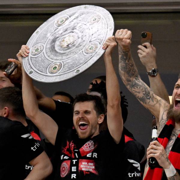 Die Leverkusener Mannschaft jubelt mit einer stilisierten Meisterschale von der Tribüne ins Stadion, den Fans zu, die nach dem Sieg das Spielfeld gestürmt haben.