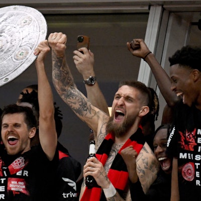 Die Leverkusener Mannschaft jubelt mit einer stilisierten Meisterschale von der Tribüne ins Stadion, den Fans zu, die nach dem Sieg das Spielfeld gestürmt haben.
