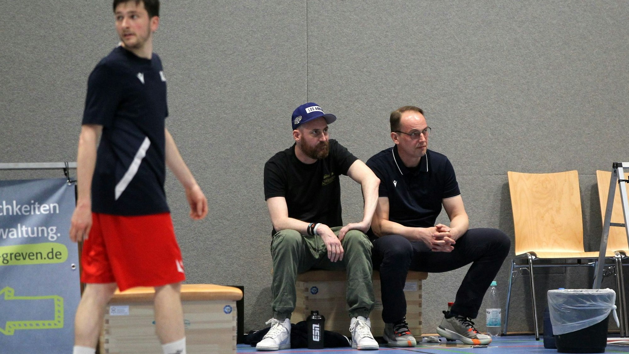 Das Trainergespann der Erftbaskets sitzt nebeneinander. Im Vordergrund ein Spieler, neben ihnen ein Mülleimer.
