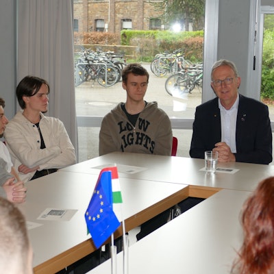 Jugendliche und ein Mann mit grauen Haaren sitzen an einem Tisch, auf dem ein EU-Fähnchen steht.