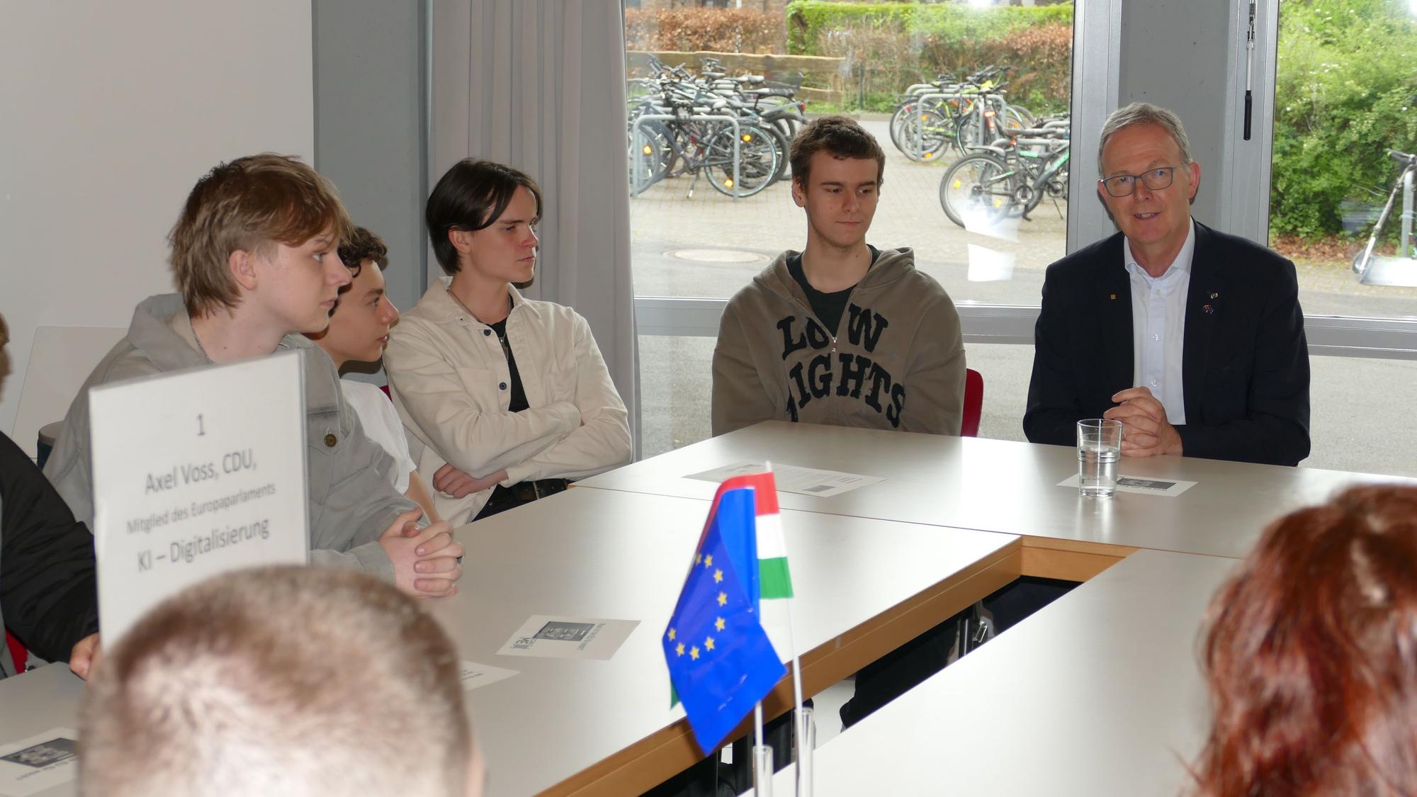 Jugendliche und ein Mann mit grauen Haaren sitzen an einem Tisch, auf dem ein EU-Fähnchen steht.