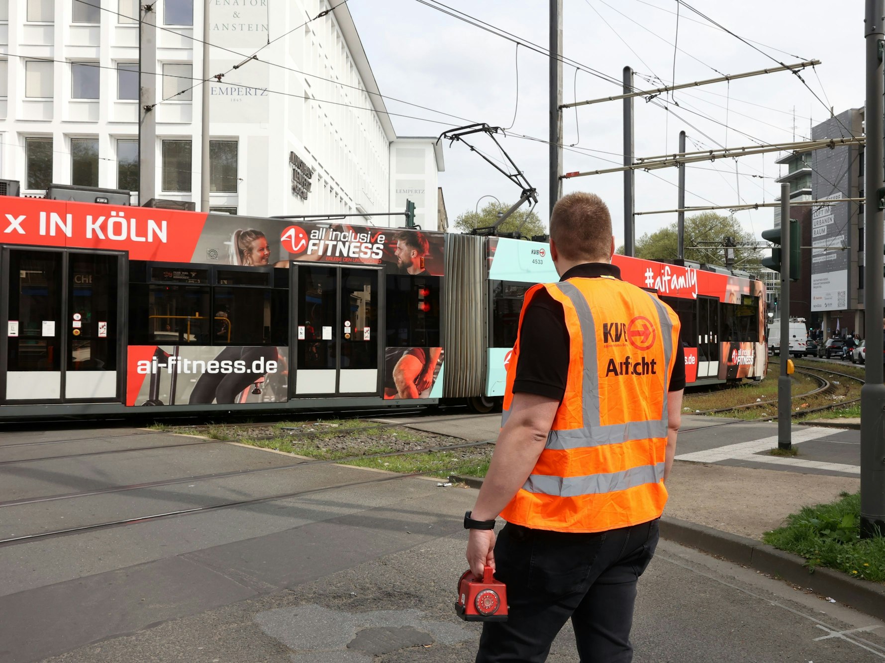 Ein KVB-Mitarbeiter mit orangefarbener Weste steht auf der Straße und schaut auf eine vorbeifahrende Straßenbahn.