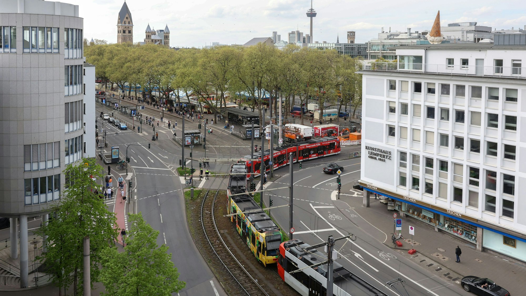 Eine aus drei Fahrzeugen bestehende, 90 Meter lange Straßenbahn fährt von der Cäcilienstraße auf den Neumarkt. Zu sehen sind die zweispurige Fahrbahn zu beiden Seiten der Gleistrasse sowie Teile des Neumarkts.
