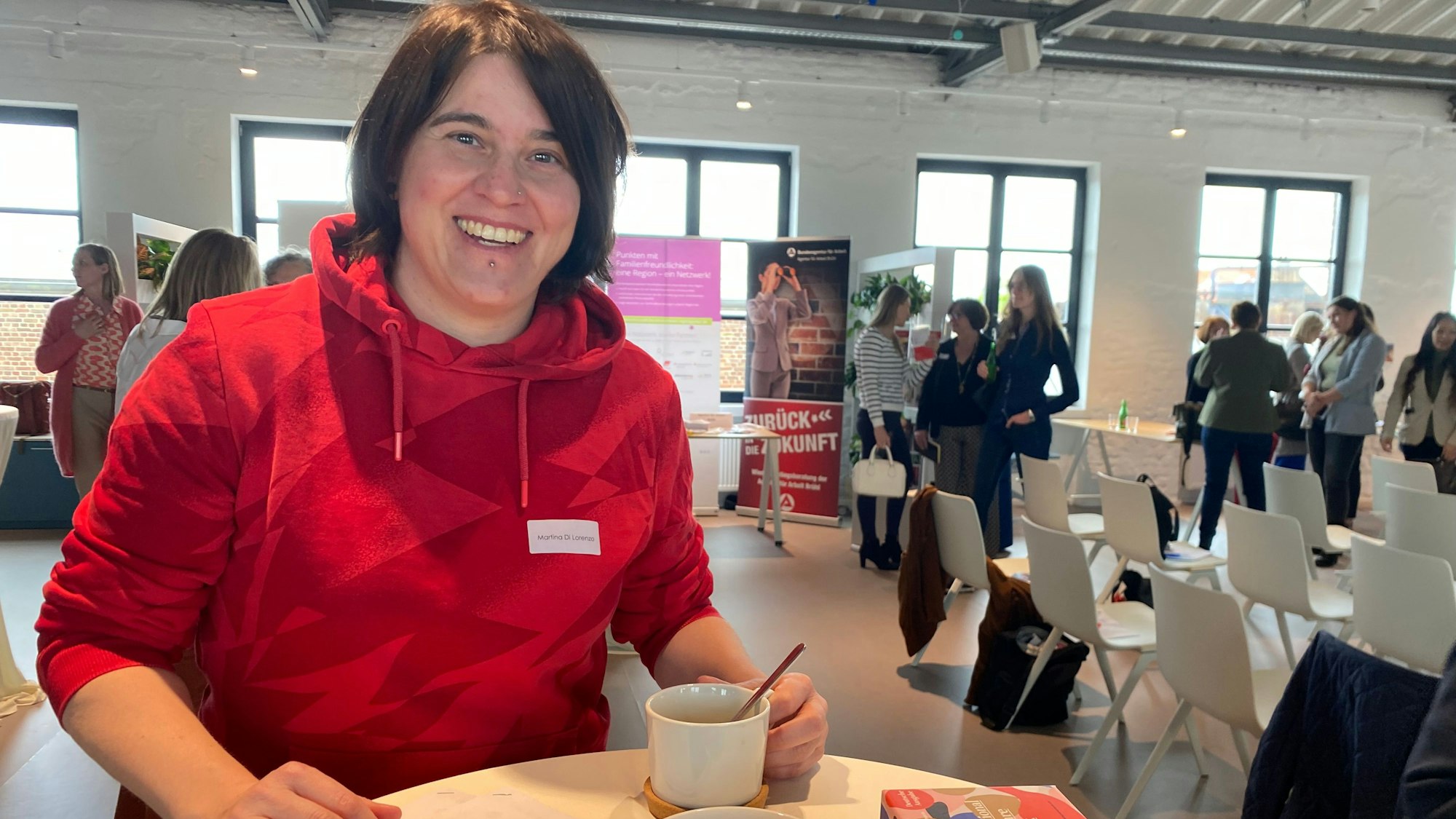 Martina Di Lorenzo steht mit einer Kaffeetasse in der Hand an einem runden Tisch. Im Hintergrund unterhalten sich andere Frauen.
