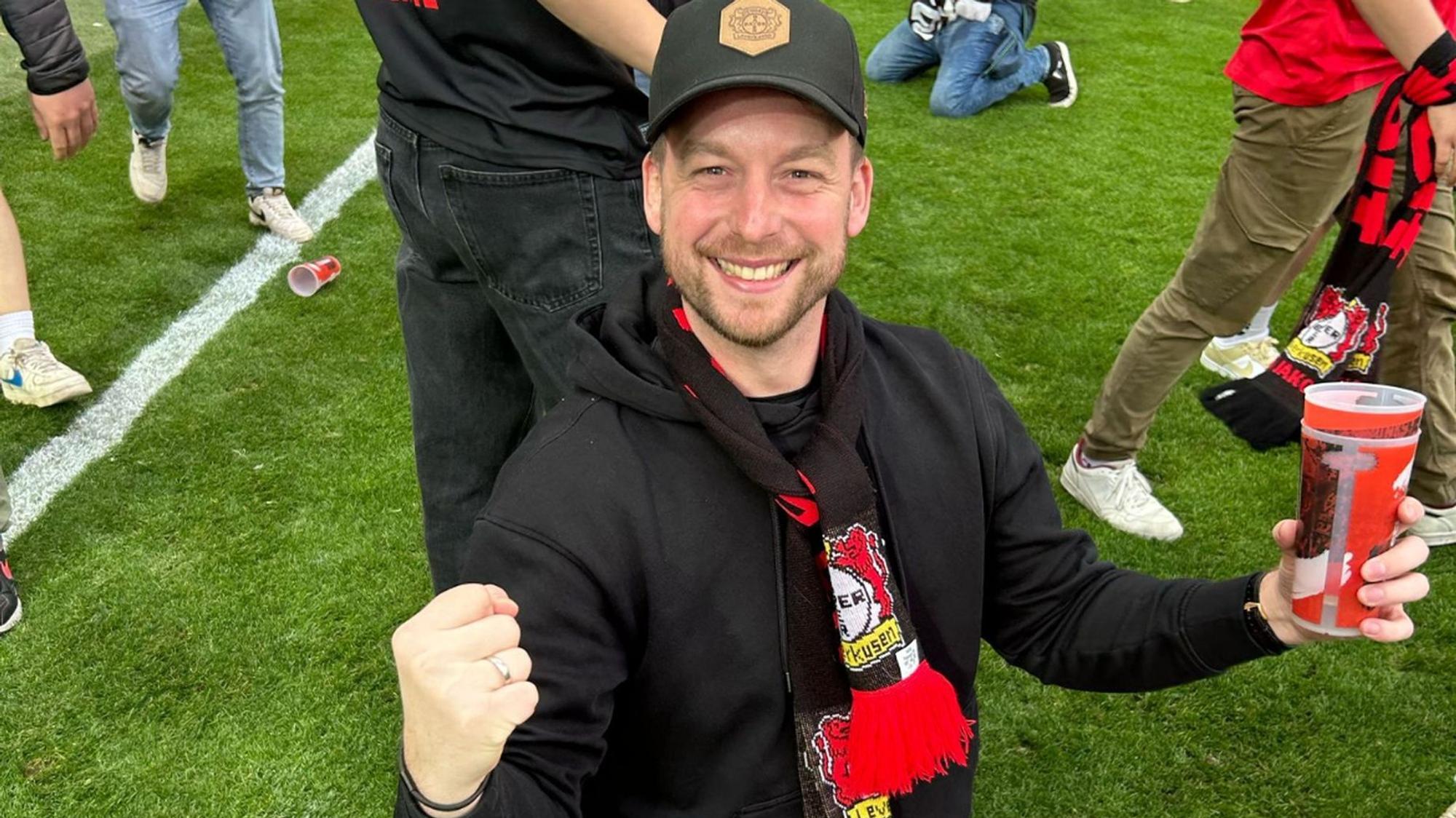 Ein Mann mit Fan-Schal von Bayer 04 Leverkusen strahlt und reckt die Faust in die Höhe. Er steht auf dem Spielfeld.
