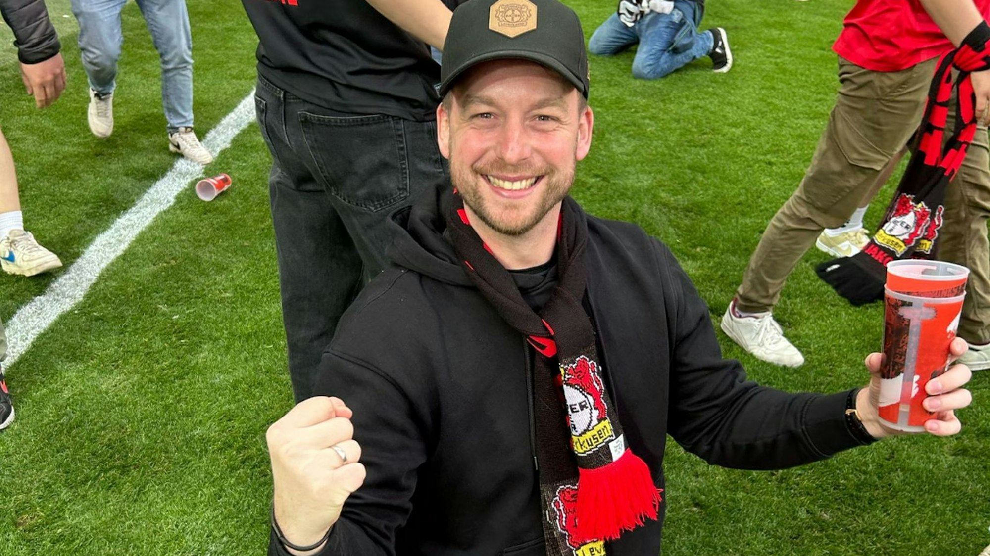 Ein Mann mit Fan-Schal von Bayer 04 Leverkusen strahlt und reckt die Faust in die Höhe. Er steht auf dem Spielfeld.