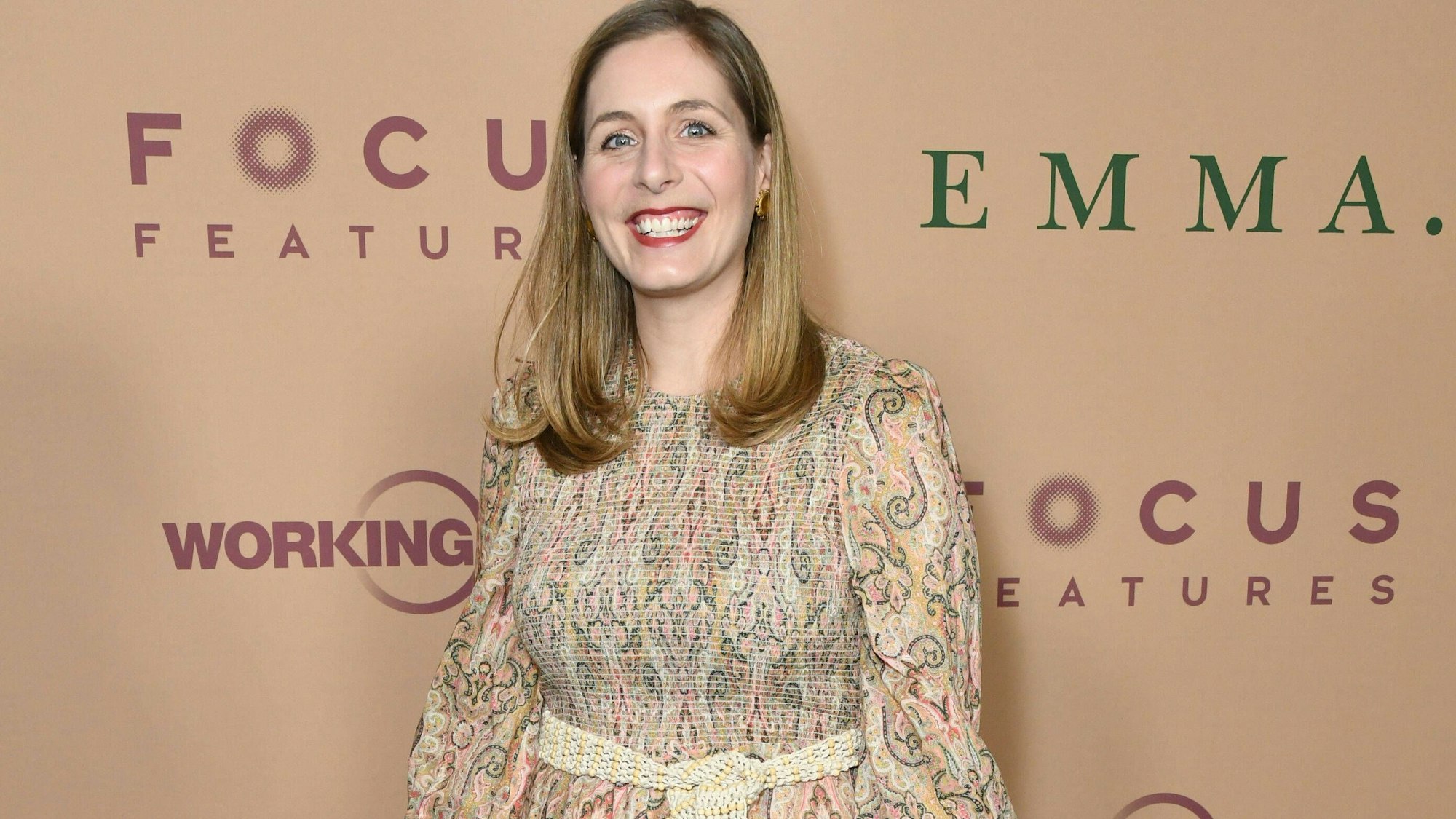 Die Autorin trägt ein gemusrertes Kleid und lächelt in die Kamera. Sie hat lange blonde Haare. Im Hintergrund eine Fotowand mit den Aufschriften "Focus Features" und "Emma".