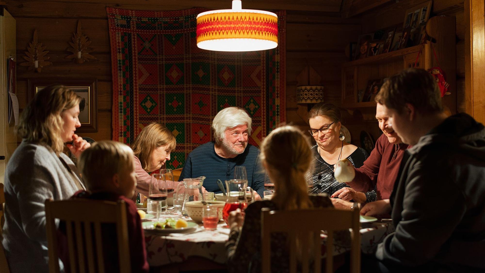 Eine Familie sitzt und speist an einem runden Essenstisch. Im Zentrum sind der Großvater, im Schatten und der Kamera den Rücken gekehrt sitzen die Kinder.