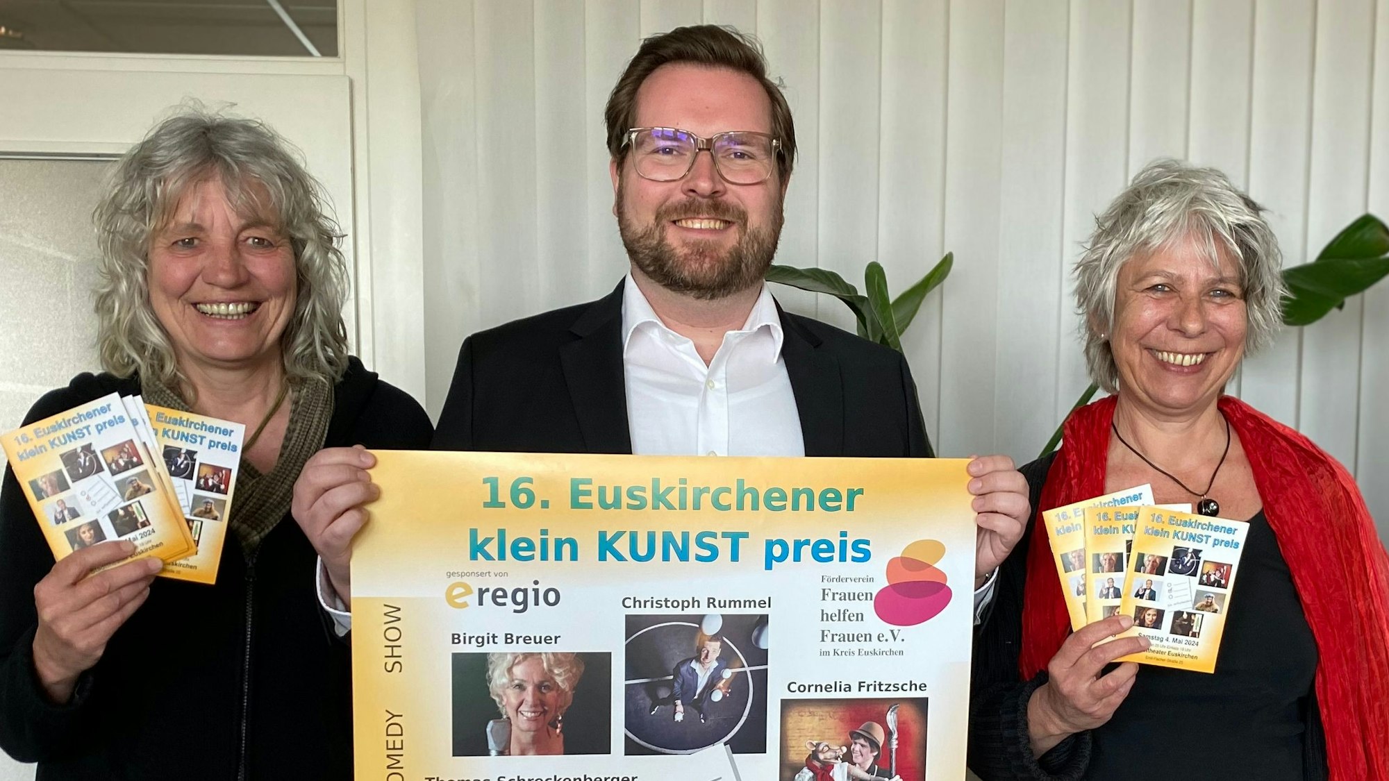 Gaby Bärenklau und Heike Jachtmann halten das Programmheft des 16. Euskirchener Kleinkunstpreises in den Händen. Sie flankieren Sebastian Zimer, der das zugehörige Plakat präsentiert.