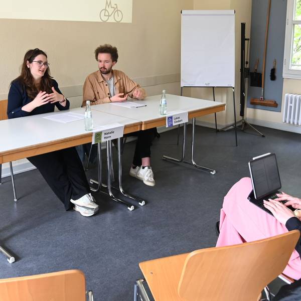 Zu sehen sind zwei Frauen und ein Mann, die auf Stühlen in einem Raum sitzen.