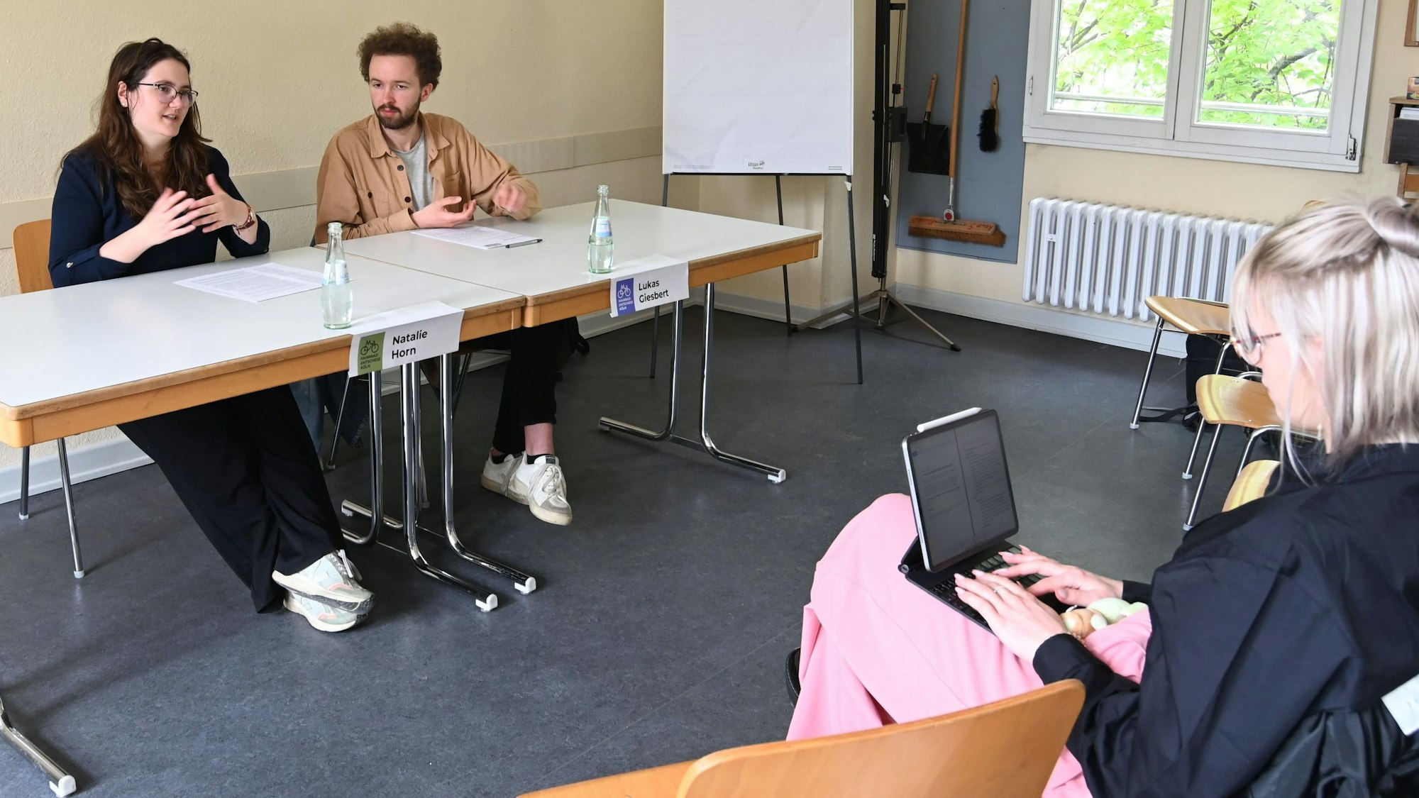 Zu sehen sind zwei Frauen und ein Mann, die auf Stühlen in einem Raum sitzen.