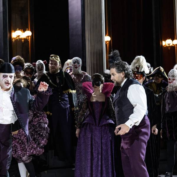Riccardo wirkt mit weiß geschminktem Gesicht und violettem Anzug ein bisschen wie der Joker. Neben ihm Simone del Savio, ebenfalls in violetter Hose, aber weißem Hemd und einer Weste. Im Hintergrund das bunte Treiben des Chores der Kölner Oper.