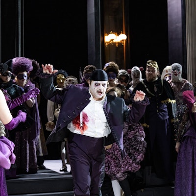 Riccardo wirkt mit weiß geschminktem Gesicht und violettem Anzug ein bisschen wie der Joker. Neben ihm Simone del Savio, ebenfalls in violetter Hose, aber weißem Hemd und einer Weste. Im Hintergrund das bunte Treiben des Chores der Kölner Oper.