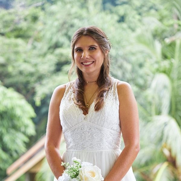 Eine lächelnde Frau mit braunen Haaren im Brautkleid. Im Hintergrund grüne Bäume.
