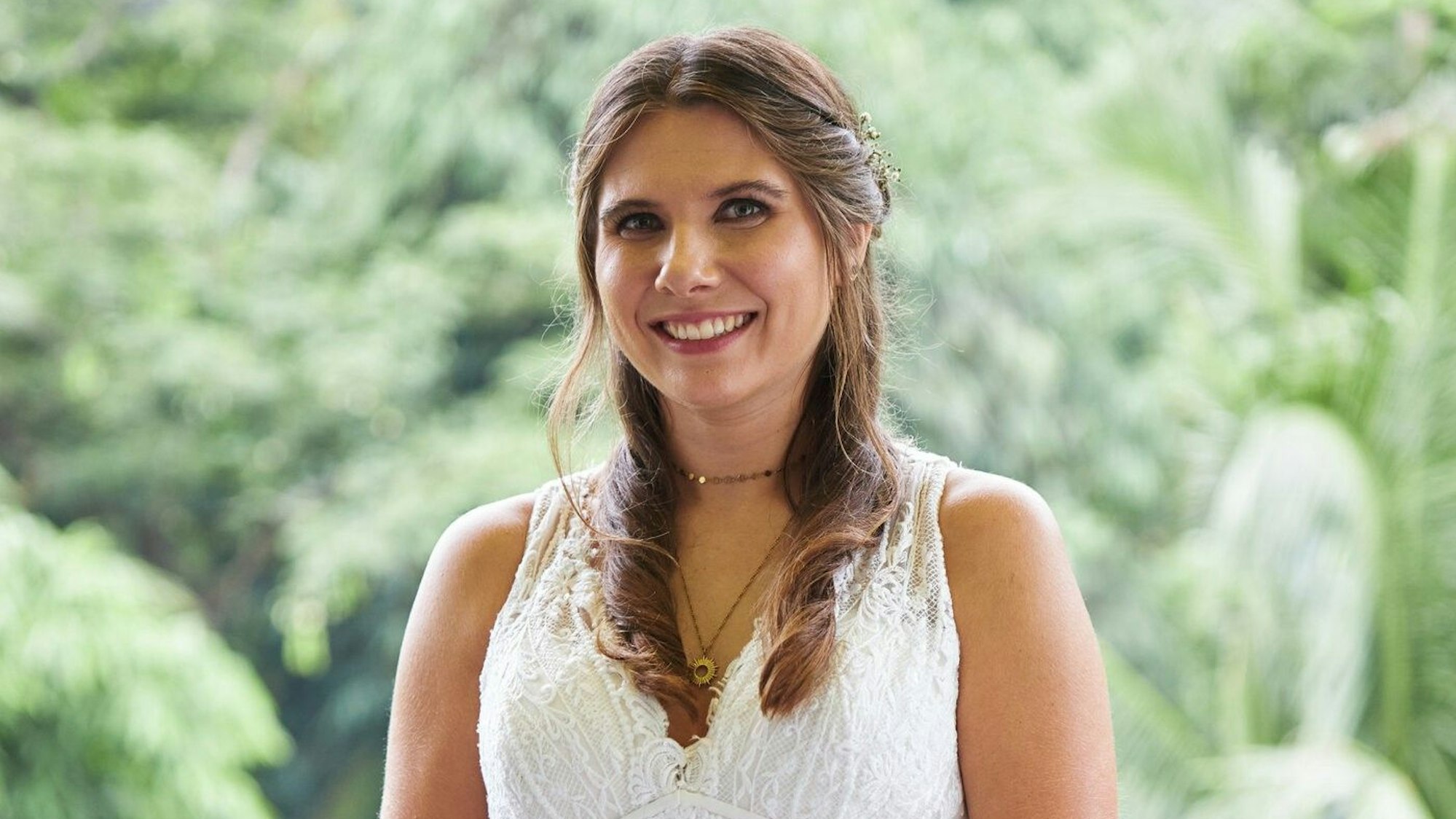 Eine lächelnde Frau mit braunen Haaren im Brautkleid. Im Hintergrund grüne Bäume.