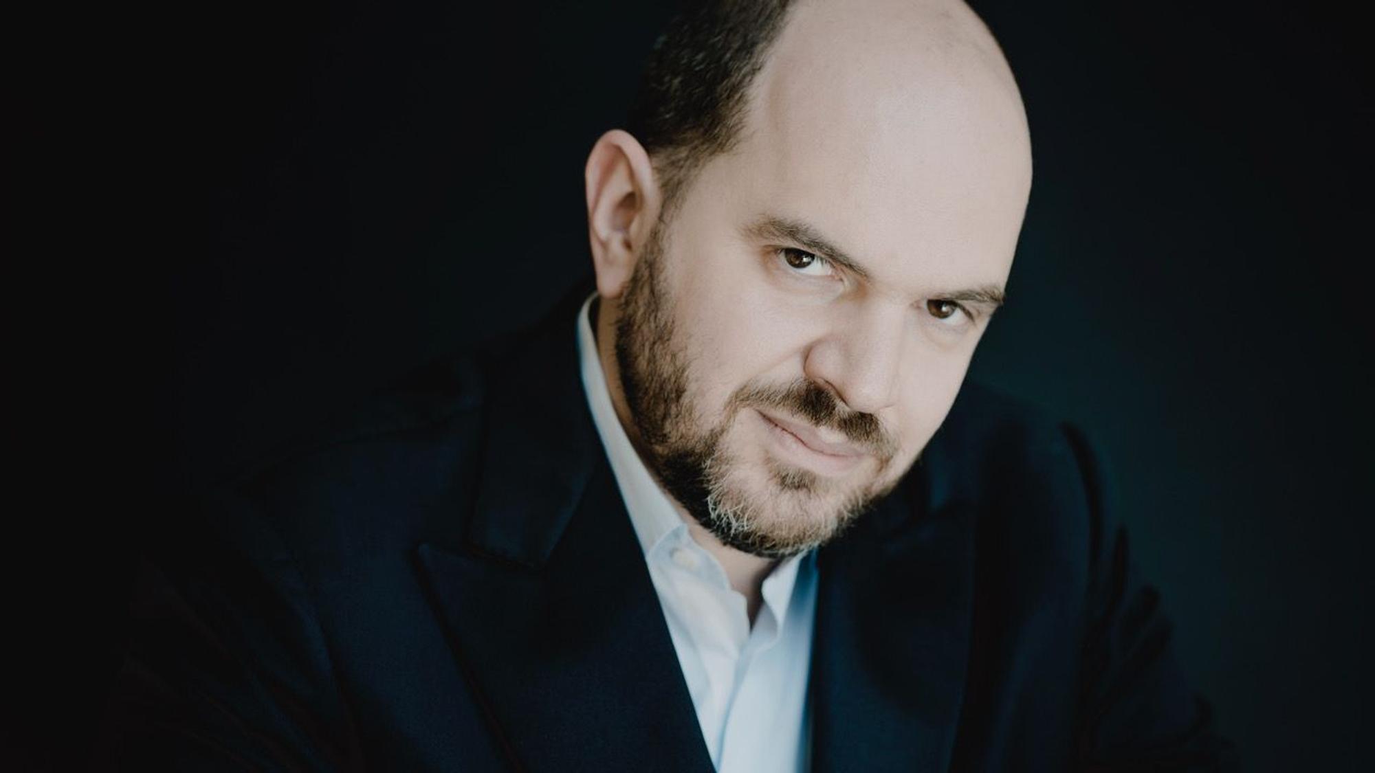 Auf dem Bild ist der Pianist Kirill Gerstein zu sehen. Er trägt einen grau melierten Bart und eine Halbglatze. Er trägt einen dunklen Anzug und ein hellblaues Hemd.