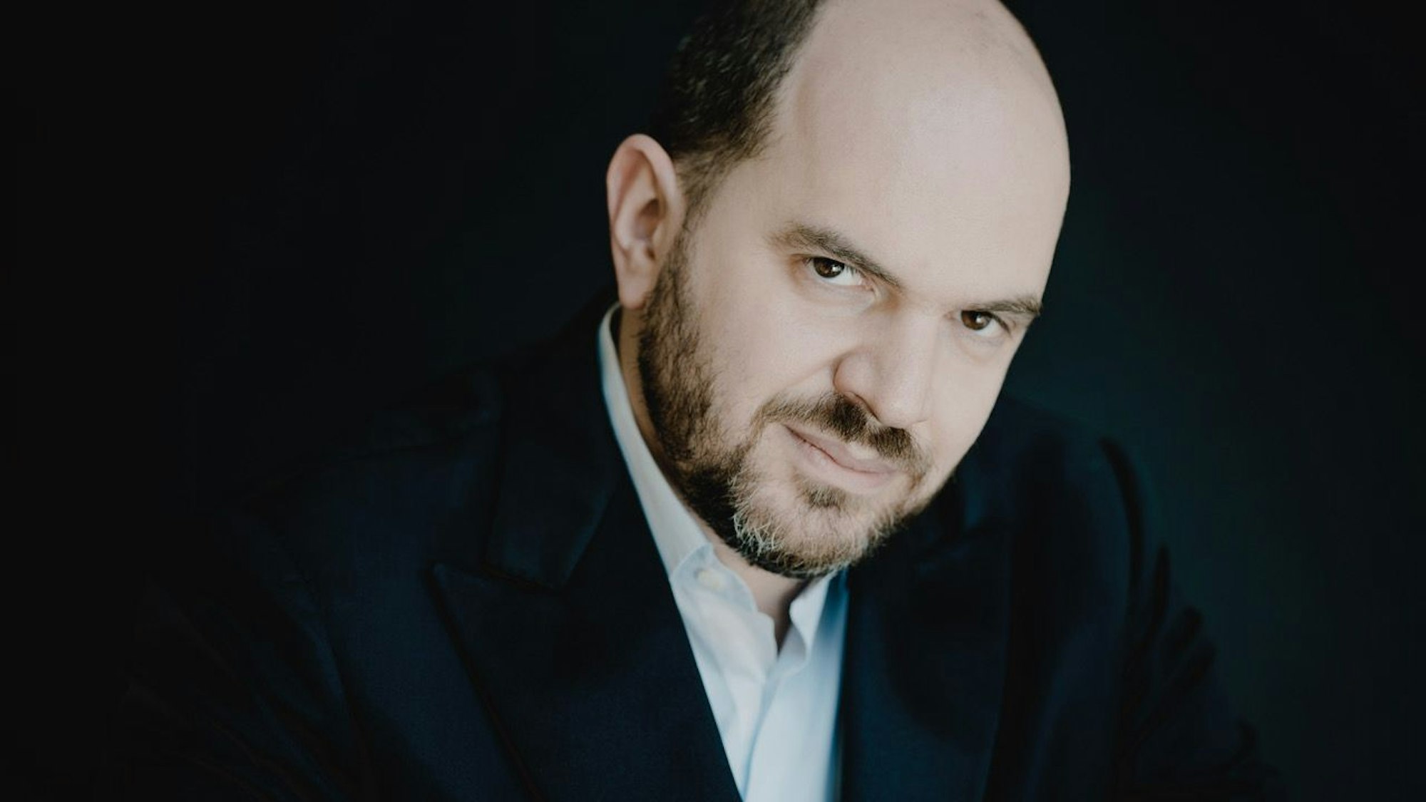 Auf dem Bild ist der Pianist Kirill Gerstein zu sehen. Er trägt einen grau melierten Bart und eine Halbglatze. Er trägt einen dunklen Anzug und ein hellblaues Hemd.