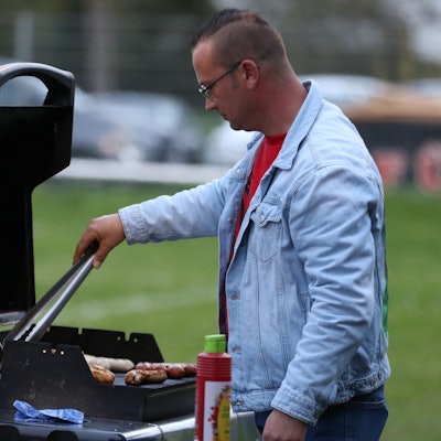 Das Bild zeigt einen Gasgrill. Ein Mann vor dem Grill dreht gerade die Bratwürstchen um.