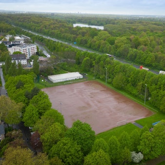Das Luftbild zeigt einen Aschenplatz inmitten von Grün zwischen Autobahn und Wohnbebauung.