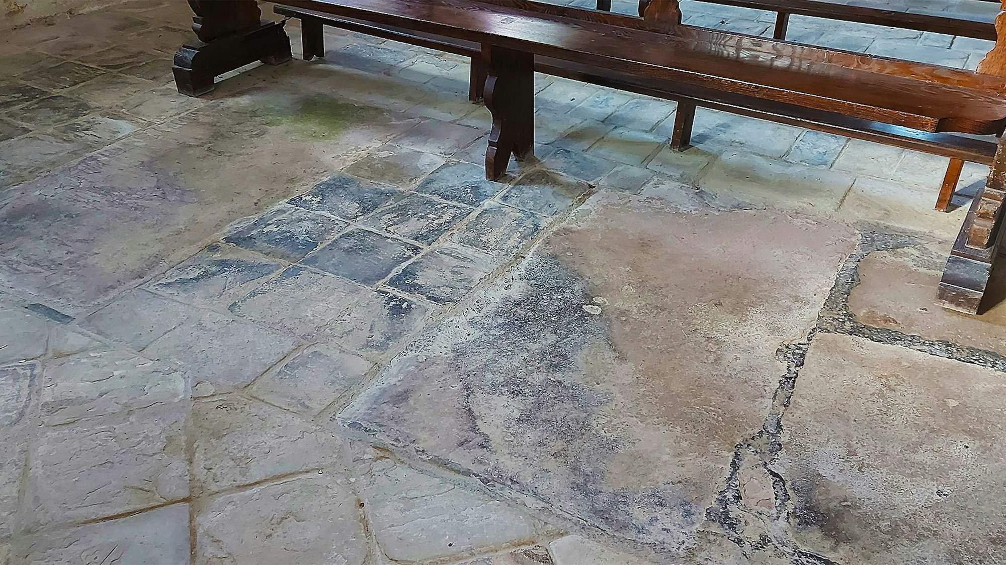 Zwei Grabplatten sind zu sehen, getrennt durch wesentlich kleinere quadratische Sandsteinplatten. Im Hintergrund steht eine Kirchenbank.