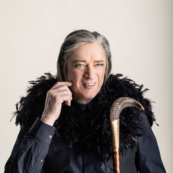 Blixa Bargeld trägt einen Schulterumhang aus schwarzen Federn und goldenen Lidschatten.