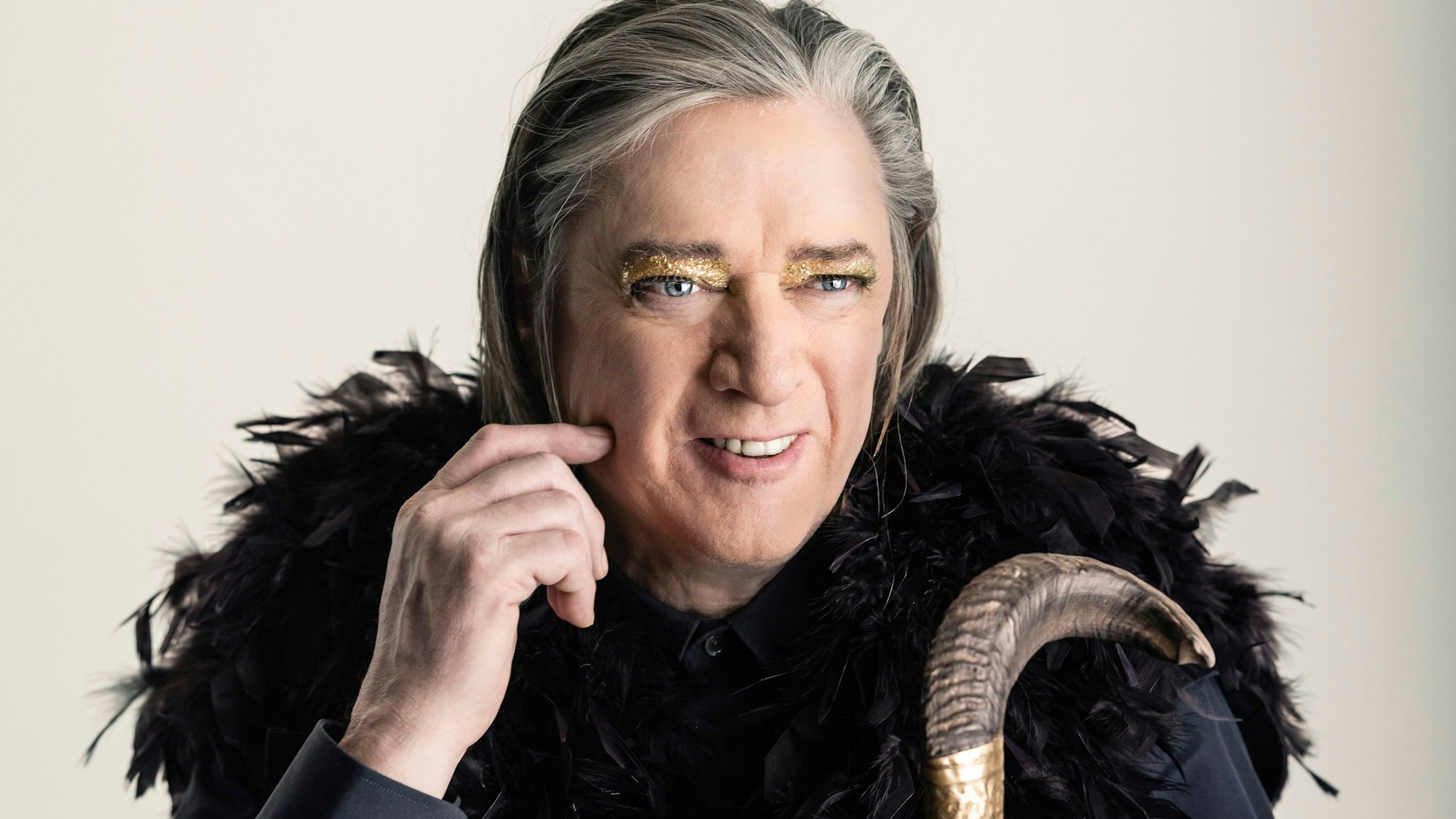 Blixa Bargeld trägt einen Schulterumhang aus schwarzen Federn und goldenen Lidschatten.