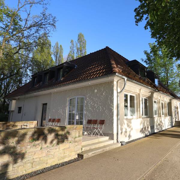 Ein flaches Gebäude mit einer Terrasse, rechts steht am Rheinufer ein Baum