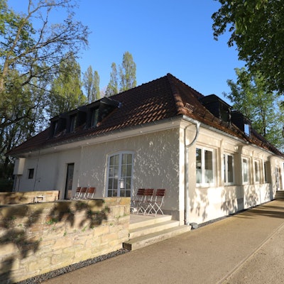 Ein flaches Gebäude mit einer Terrasse, rechts steht am Rheinufer ein Baum