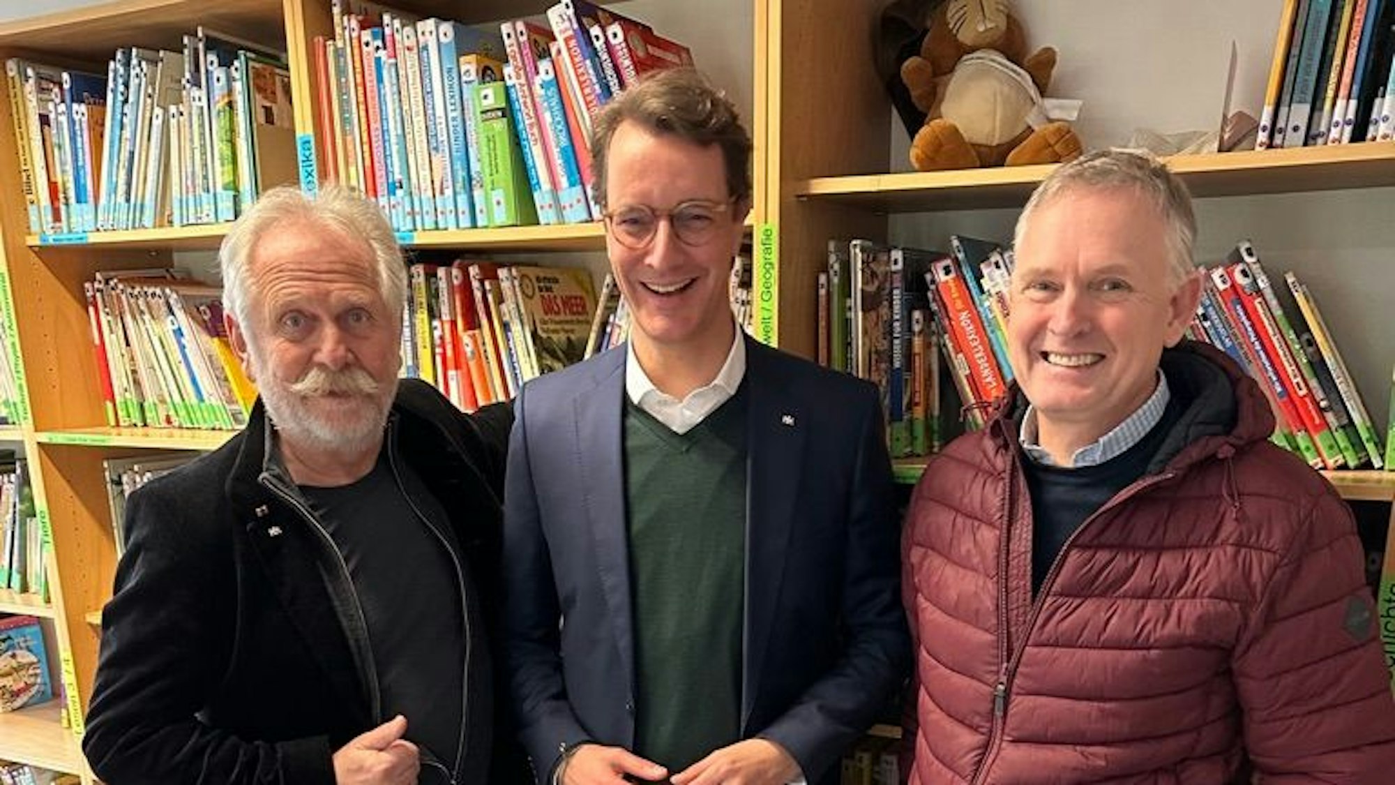 Der Kölner Musiker Henning Krautmacher, NRW-Ministerpräsident Hendrik Wüst und „Rund an Ride for Reading“-Gründer Oliver Gritz stehen vor einem Bücherregal in einem Kölner Leseclub.