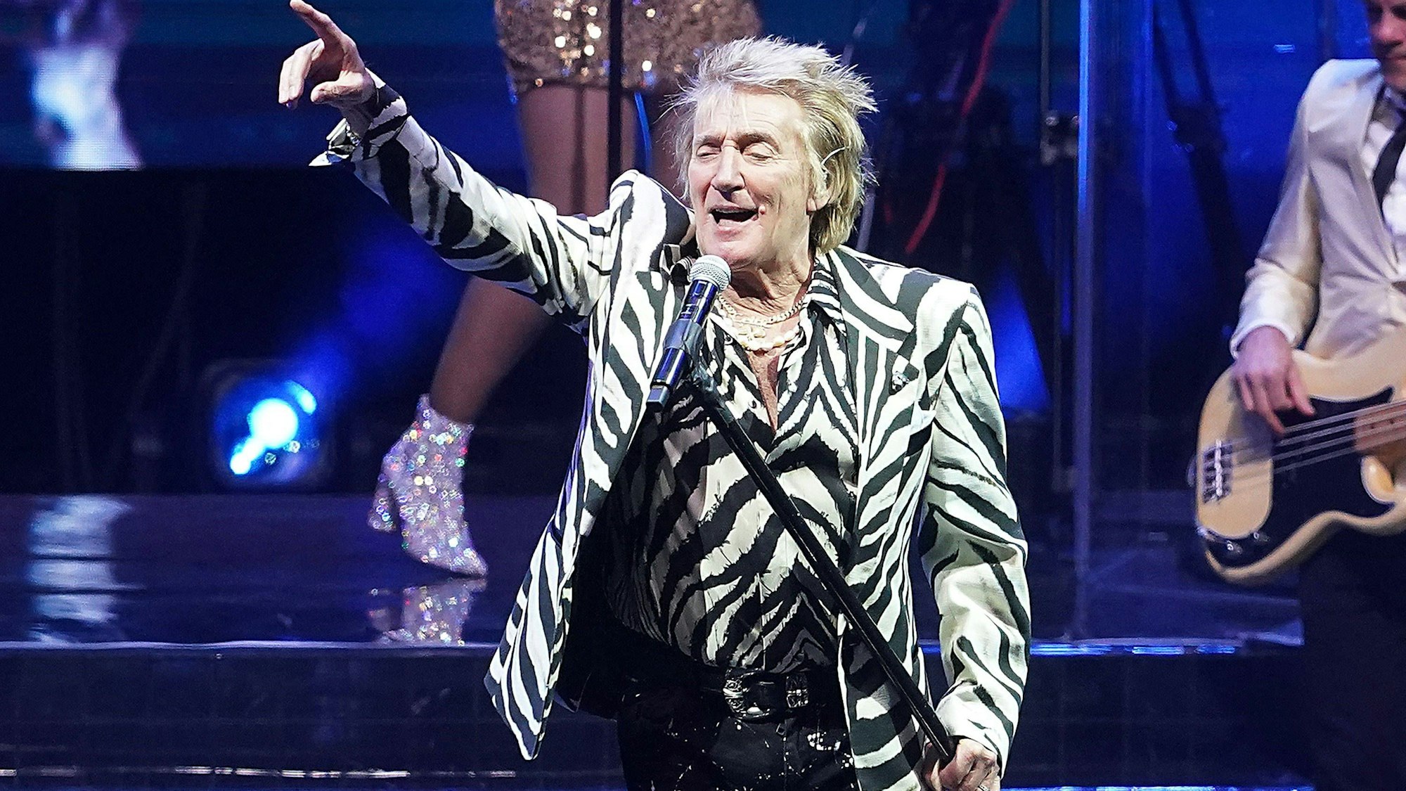 Rod Stewart trägt ein Zebra-gemustertes Sakko und ein dazu passendes Hemd. Er hält mit einer Hand die Mikrofonstange und zeigt mit der anderen nach links oben im Bild. Im Hintergrund ein Gitarrist und eine weitere Sängerin, die Bühne wird bläulich erleuchtet.