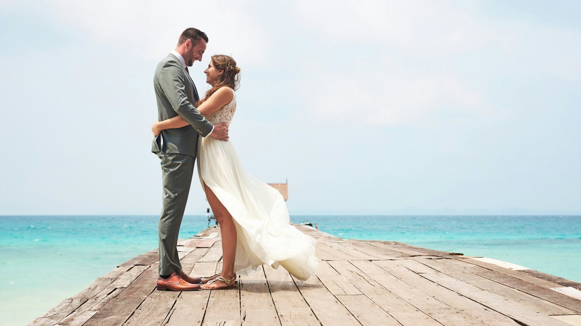 Saskia und Jörn in ihrer Hochzeitsgarderobe auf einem Steg, der ins blaue Meer führt.