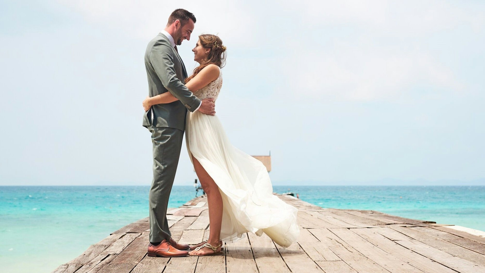 Zwei Personen in festlicher Kleidung stehen auf einem Steg. Hinter ihnen blaues Wasser und blauer Himmel.