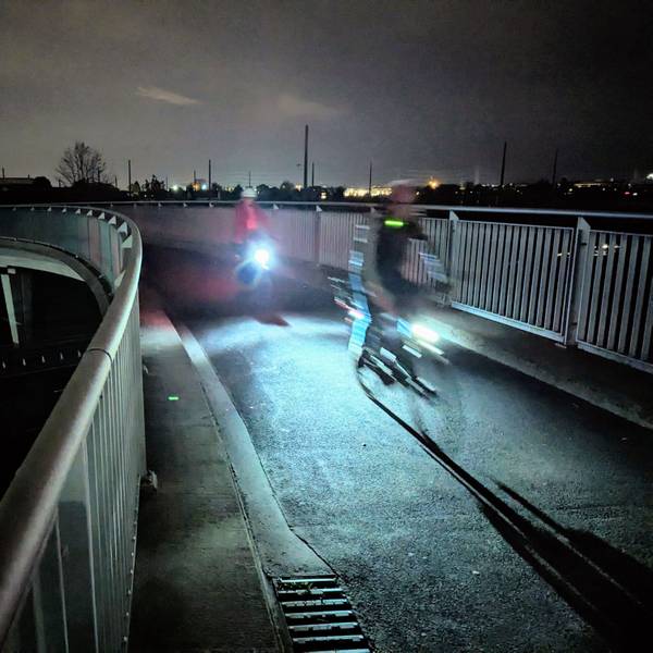 Zwei Radfahrer mit beleuchteten Fahrrädern fahren in der Dunkelheit über eine Brücke.