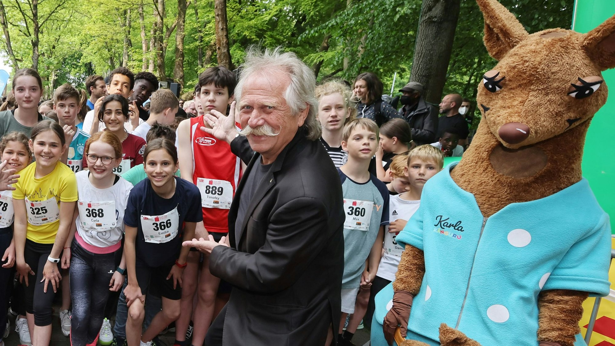 Musiker und Ey-Höhner-Frontmann Henning Krautmacher steht am Startpunkt des Laufs der Schulen und zeigt auf Schülerinnen und Schüler, die eine Startnummer tragen.