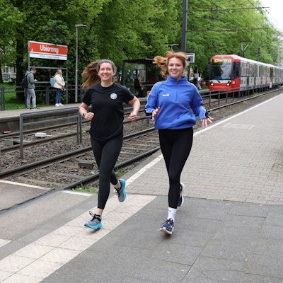Die Kölnerinnen Katharina Oeltermann und Sophia Staudinger tragen Sportkleidung und laufen an der KVB-Haltestelle Ubierring entlang.
Foto von Martina Goyert