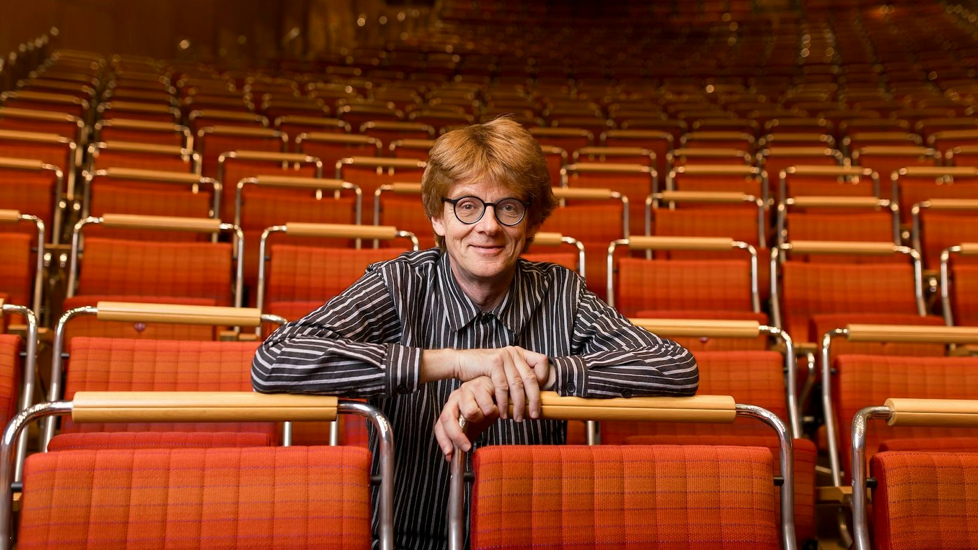 Enno Poppe sitzt in einer Stuhlreihe in der Kölner Philharmonie und lehnt mit den Armen auf den Rückenlehnen der Vorderreihe.