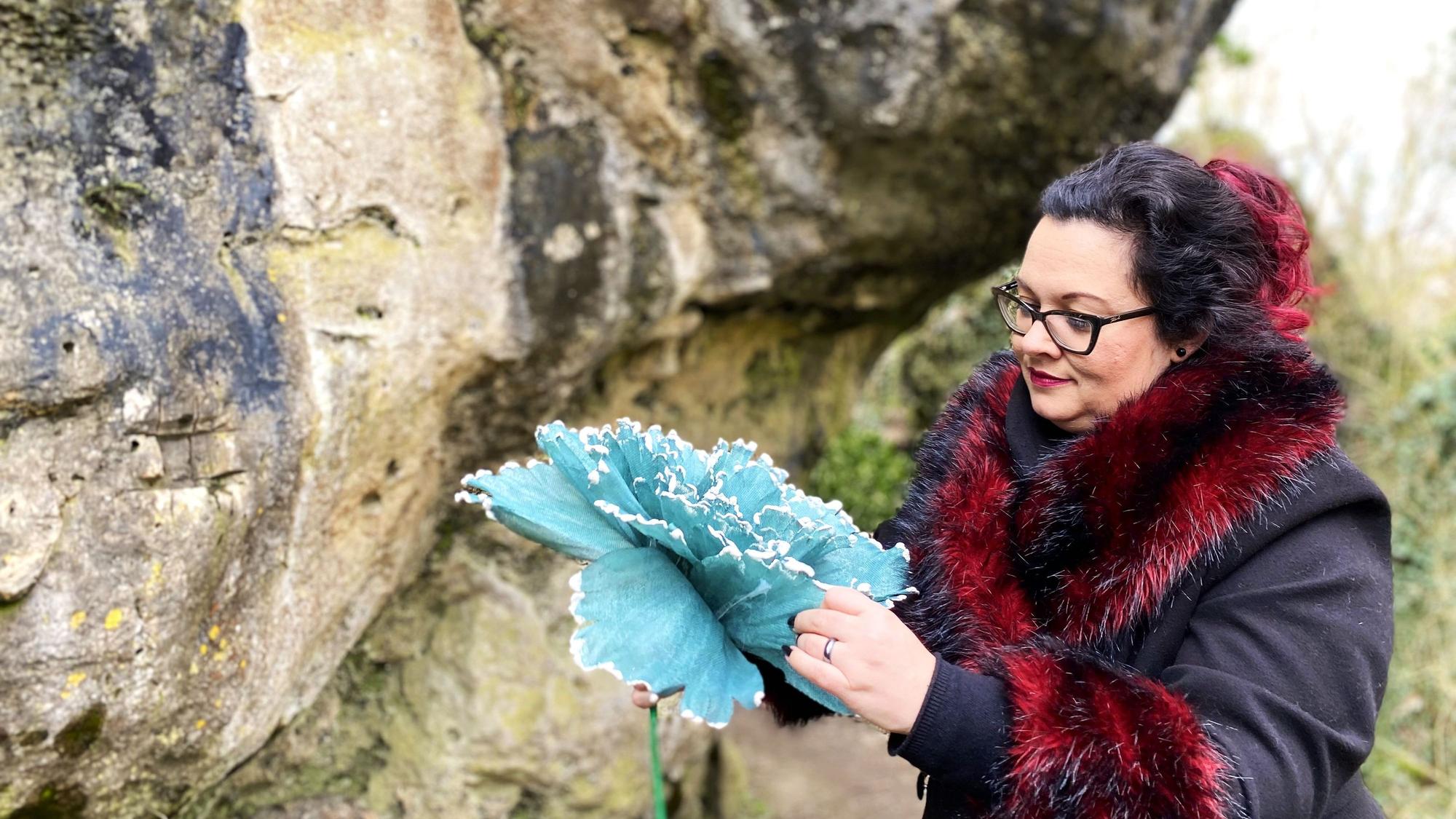 Vanessa Gebhardt steht vor der Kakushöhle in Mechernich und faltet eine selbstgebastelte blaue Blume auf.