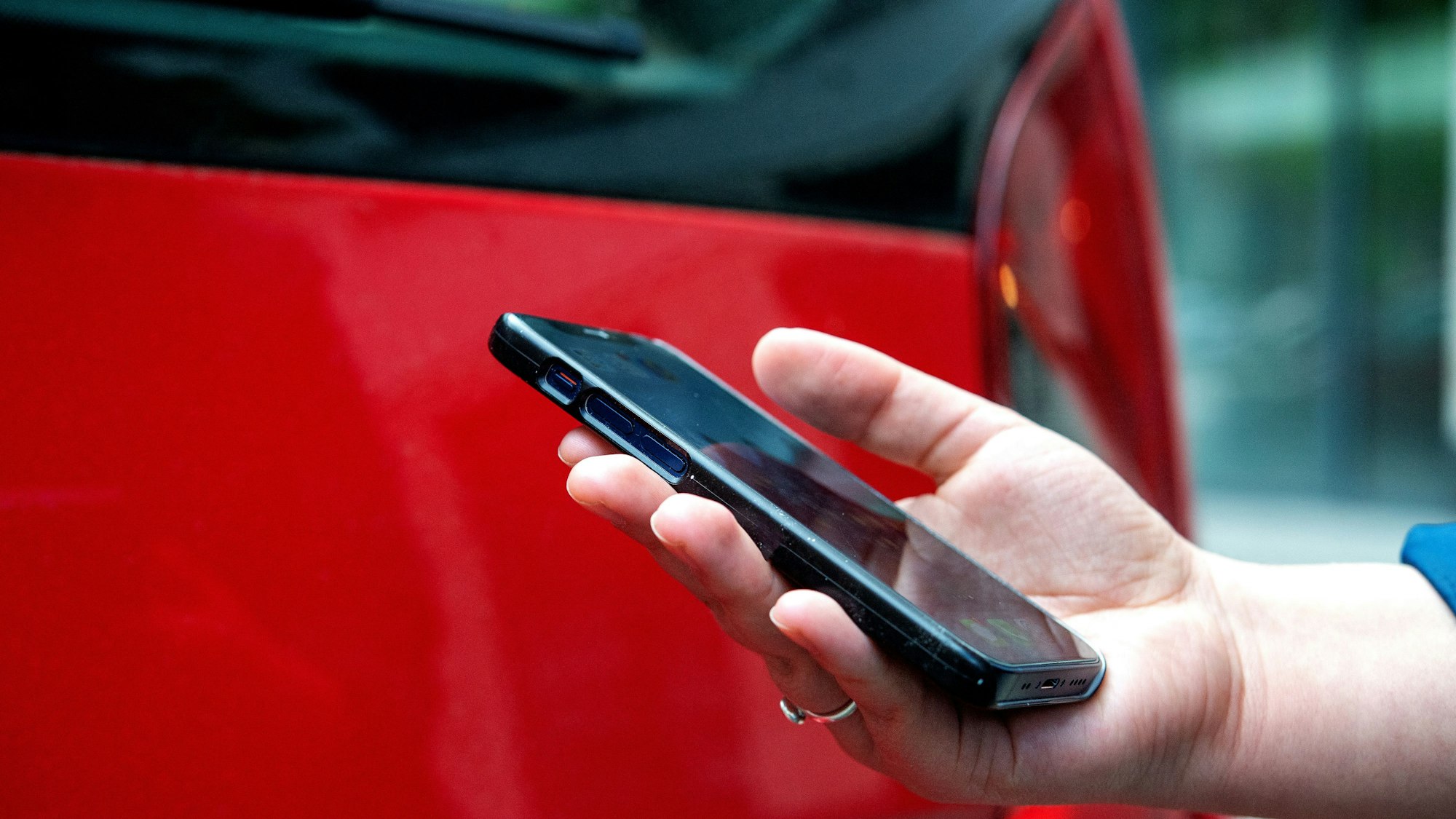 Das Foto zeigt eine Hand mit einem Smartphone und im Hintergrund ein rotes Auto.