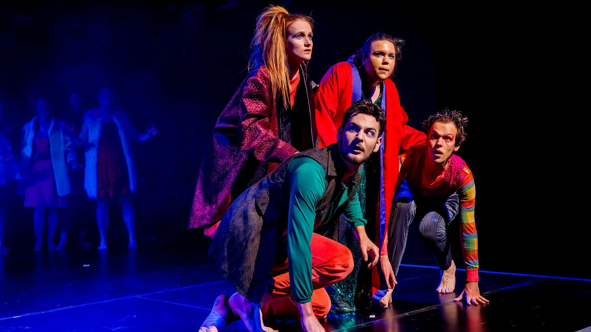 Zu sehen sind zwei hockende Männer und dahinter zwei Frauen, die sich auf sie stützen. Alle tragen bunte Kleidung. Der Hintergrund ist schwarz, auf einer Seite ist die Bühne blau beleuchtet.