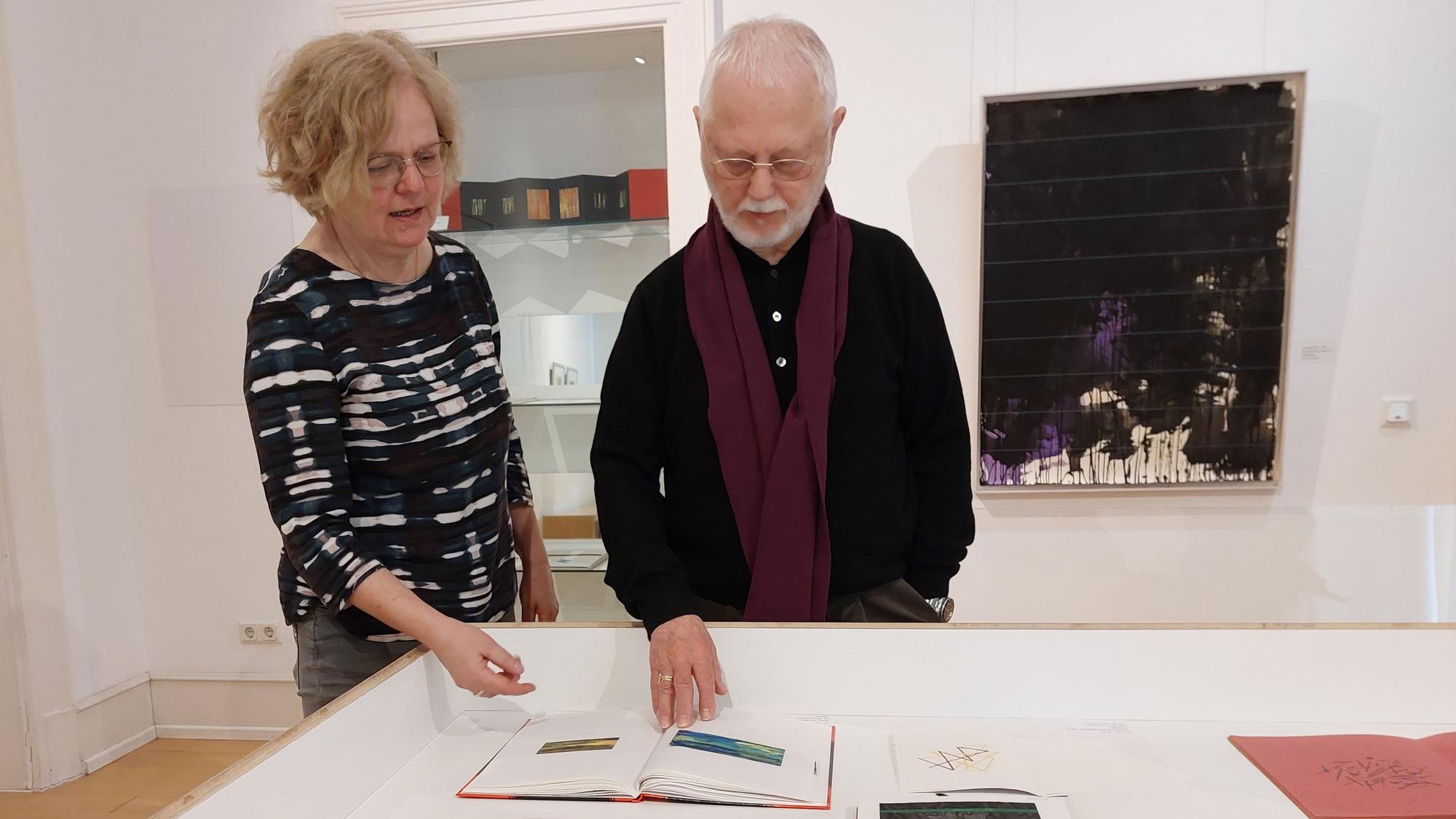 Eine blonde Frau mit Brille und ein älterer Herr betrachten ein Buch in einer Vitrine.