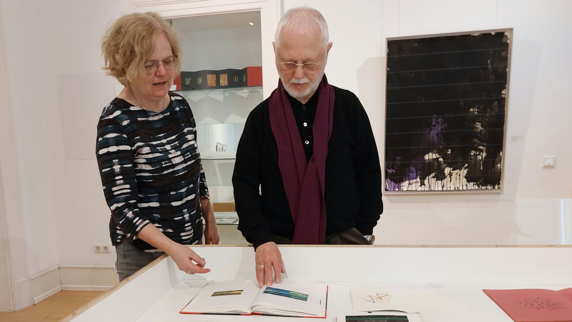 Eine blonde Frau mit Brille und ein älterer Herr betrachten ein Buch in einer Vitrine.
