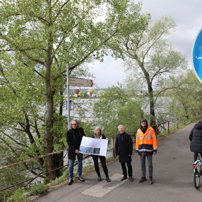 Eine vierköpfige Gruppe steht auf einem Radweg, am Rand weisen rot-weiße Schilder nach Bonn und Königswinter..