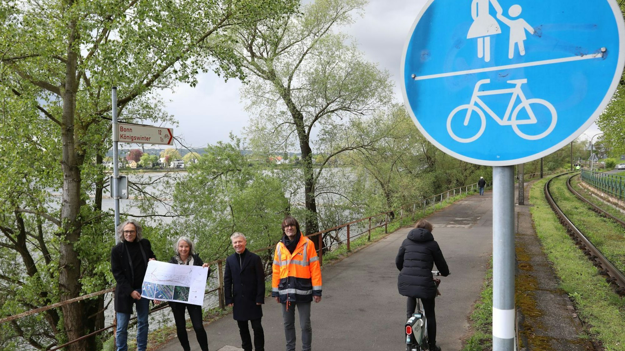 Eine vierköpfige Gruppe steht auf einem Radweg, am Rand weisen rot-weiße Schilder nach Bonn und Königswinter..