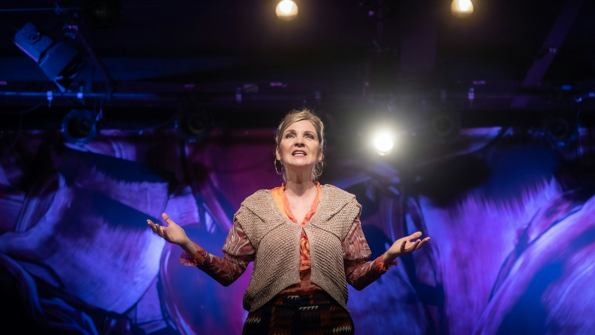 Susanne Pätzold trägt eine braune Weste und darunter ein oranges, gemustertes Top. Sie hat blonde Haare und trägt Reifenohrringe. Der Hintergrund ist abstrakt und bläulich-violett. Man sieht über der Bühne ein paar Bühnenscheinwerfer.