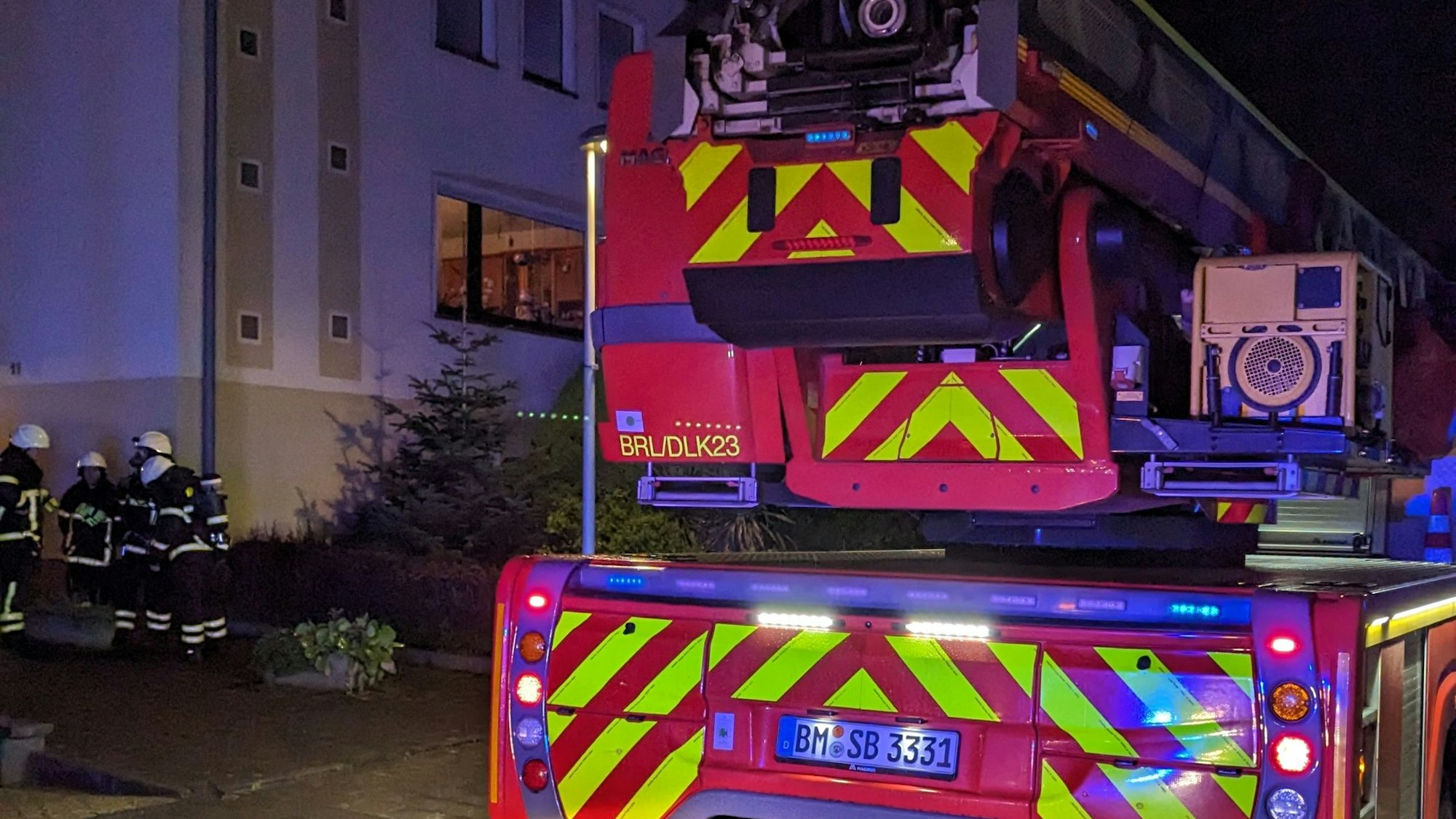 Ein Feuerwehreinsatzfahrzeug. Im Hintergrund Einsatzkräfte und ein Mietshaus.