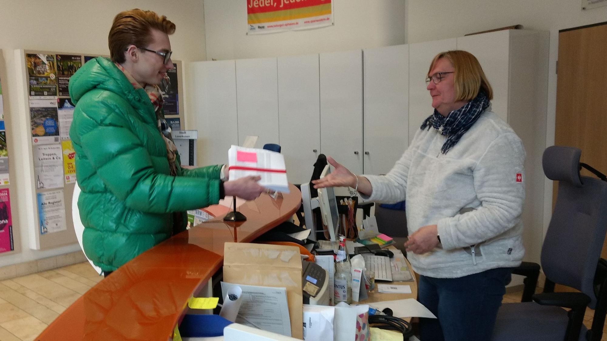 Ein Mann übergibt einer Frau im Foyer des Rathauses Leverkusen einen Stapel Papier.
