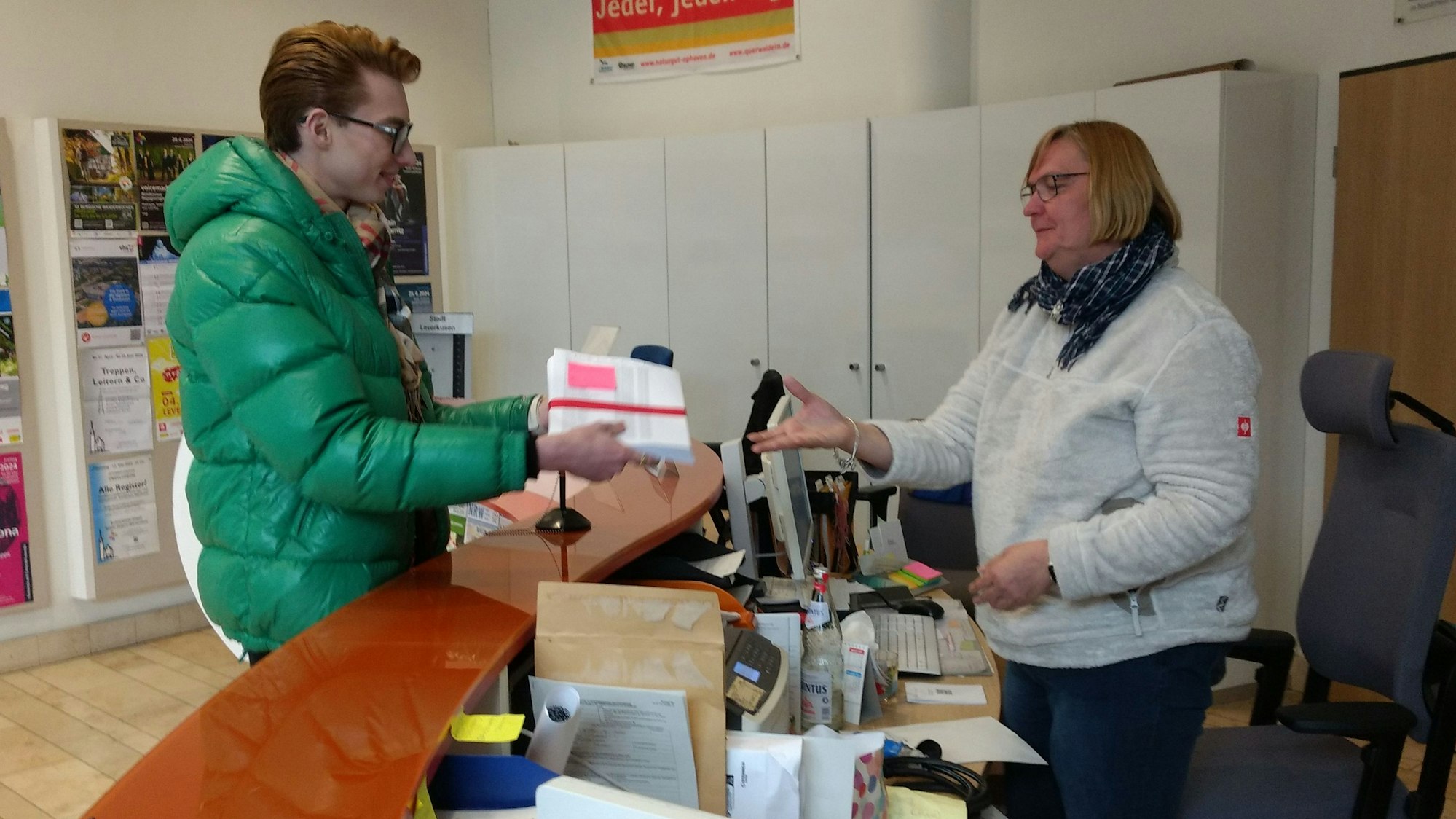 Ein Mann übergibt einer Frau im Foyer des Rathauses Leverkusen einen Stapel Papier.