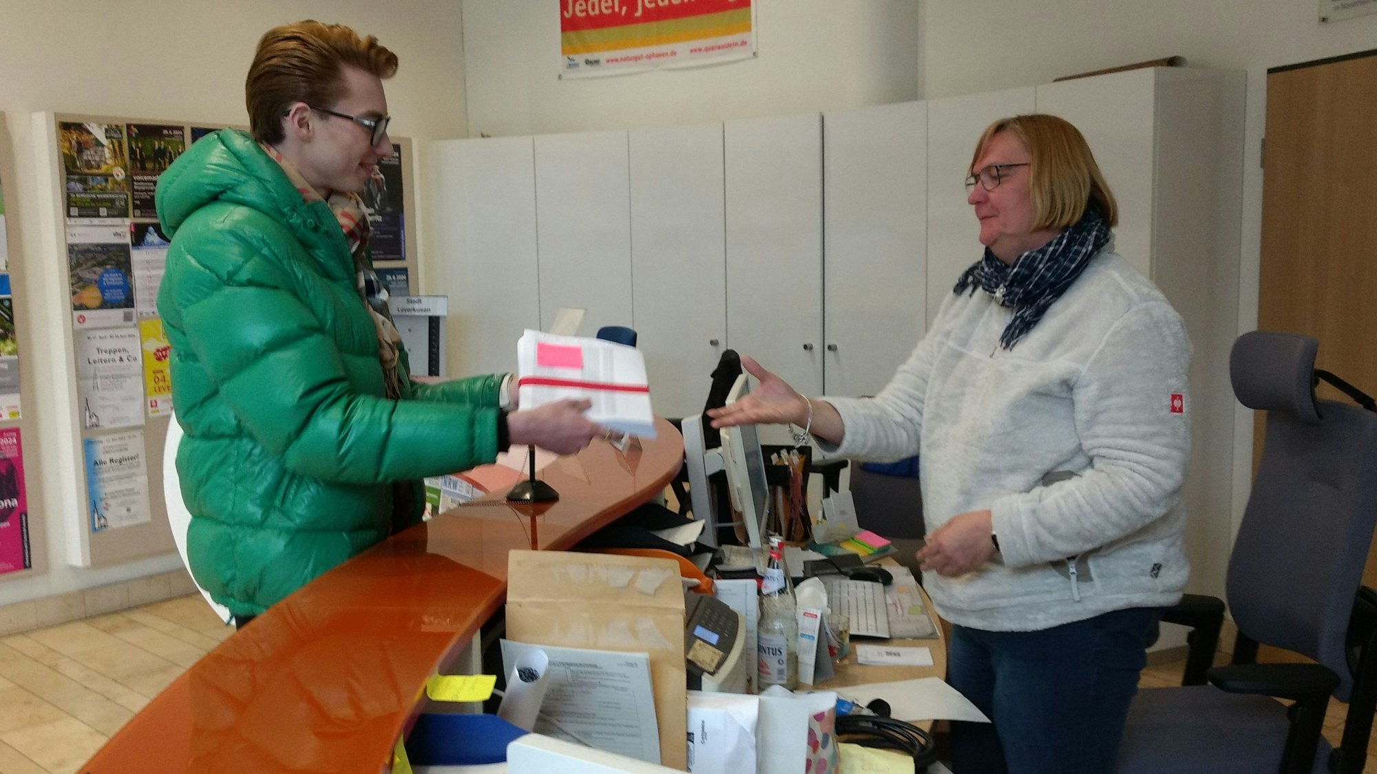 Ein Mann übergibt einer Frau im Foyer des Rathauses Leverkusen einen Stapel Papier.
