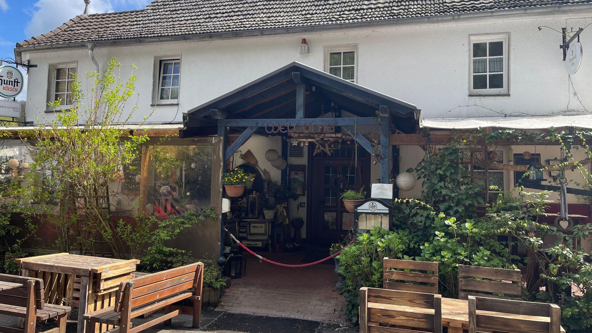 Vier Personen sitzen an einem Tisch im Freien. Im Hintergrund steht ein Gebäude mit einem Schild „Bauernschänke Bröleck“.
