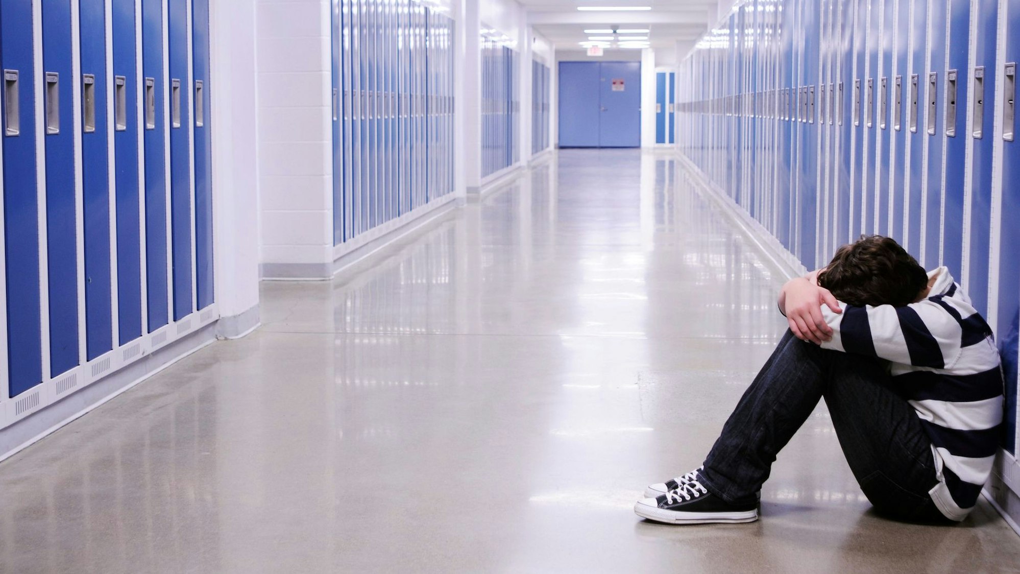 Ein Junge sitzt auf dem Boden eines Flures mit vielen blauen Spinden, er hält seinen Kopf zwischen den verschränkten Armen.